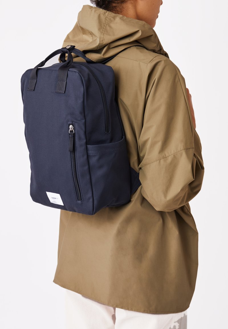 Laadukkaasta tummansinisestä kankaasta valmistettu reppu, jossa on vetoketjullinen etutasku, säädettävät olkaimet ja sivutasku, kuljetettuna oliivinvihreän takin päällä.