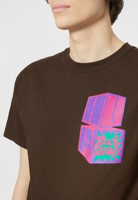 T-shirt en coton marron arborant un graphique géométrique vibrant en rose, violet et vert avec l'inscription "HU Rock Steady" sur le devant.