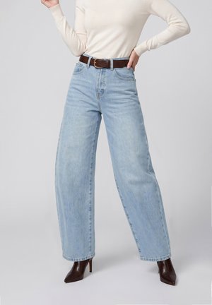 Personne portant un jean large bleu clair, une ceinture marron, un haut crème à manches longues et des bottines marron pointues à talons hauts, debout la main sur la hanche.