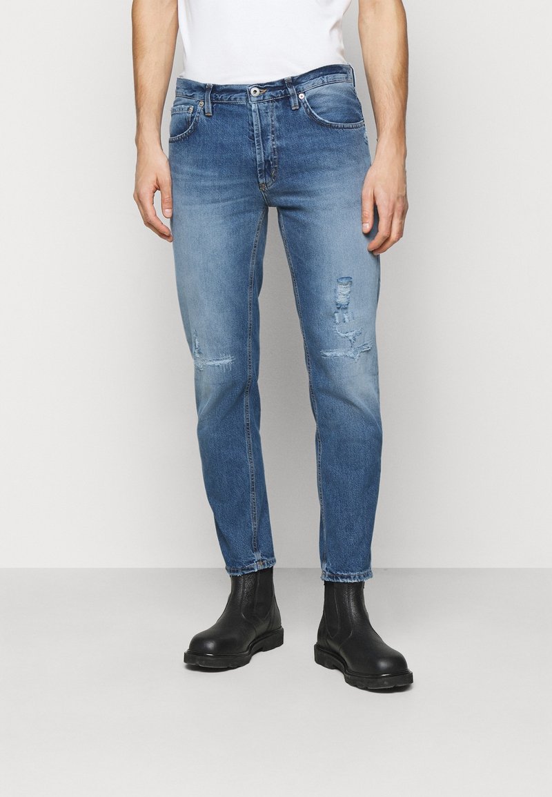 Homme portant un jean bleu délavé et des bottines en cuir noir, debout devant un fond clair uni.
