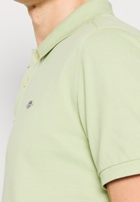 Gros plan sur une personne portant un polo vert clair avec un petit logo brodé d'un drapeau américain sur la poitrine.