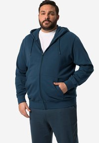 Námořnická mikina na zip s přední kapsou a kapucí na šňůrku, vyrobená z měkké tkaniny; spárovaná s odpovídajícími tmavými kalhotami.