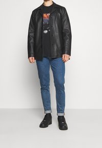 Svart läderjacka med kragdesign, med knapplås, bärs över en grå t-shirt med grafik och blå jeans. Svarta sneakers fulländar looken.