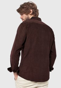 Ruskea corduroy-paita terävillä kauluksilla, pitkillä hihoilla ja napillisia mansetteja; tarjoaa väljän istuvuuden ja tekstuurin.
