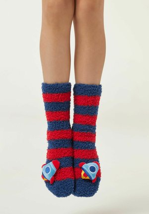 Nogi dziecka ubrane w niebiesko-czerwone paskowane, puszyste skarpetki z małymi dekoracjami w kształcie rakiety na palcach, na jednolitym tle.