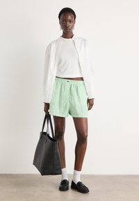 Shorts în dungi verzi și albe, top alb casual, geantă neagră tip tote și loafers negri. Modelul stă pe un fundal neutru.