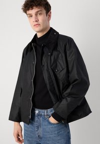 Must värvi kerge jakk koos kootud kraega, nööpide ja rinnataskuga, kaasas must turtleneck ja sinised teksad.