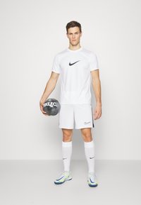 Vitt Nike sporttröja och shorts med svarta logotypdetaljer, hållande en grå Nike F.C. fotboll. Vita strumpor och flerfärgade träningsskor.