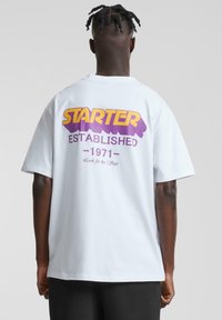 Hvit bomulls t-skjorte med stor lilla og gul grafisk tekst på baksiden som leser "STARTER ESTABLISHED - 1971" med en jevn tekstur.