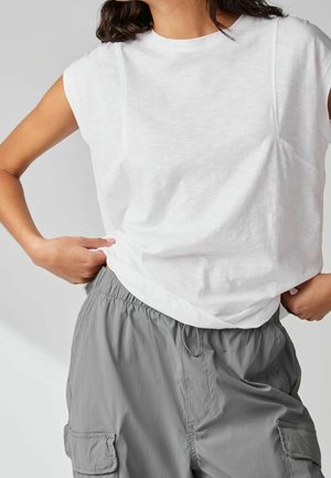 Vrouw draagt een mouwloos wit topje en losse grijze cargobroek met elastische taille, staand met handen bij de heupen.