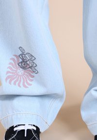 Un pantalon en denim bleu clair à coupe décontractée présente un motif graphique rose et noir sur la jambe gauche, représentant un soleil et des formes abstraites.