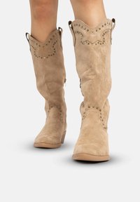 Helle Wildleder-Stiefel mit knielangem Schaft, gezacktem oberen Rand, dekorativen Nähten und Metallnieten an den Kanten. Niedriger Blockabsatz, abgerundete Spitze.