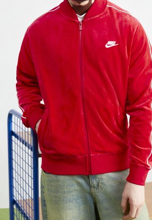 Osoba nosząca czerwoną kurtkę Nike na zamek błyskawiczny z białymi paskami na rękawach i niebieskie dżinsy, stoi z jedną ręką w kieszeni.