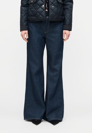 Person iført mørkeblå vidde bukser i denim, sort quiltet jakke og sorte spidse sko, stående mod en ensfarvet baggrund.