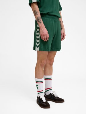 Osoba ubrana w ciemnozielone szorty z białymi skośnymi paskami po bokach, białe skarpetki z czerwonymi i zielonymi paskami oraz czarne trampki, stojąca na białym tle.