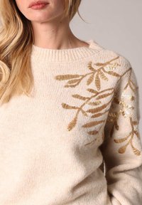 Crème gebreide trui met gouden pailletten bloemenontwerp op de linkerschouder en mouw, met een geribbelde halslijn en zachte textuur.