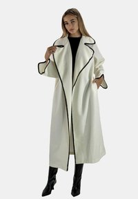 Lang hvid frakke med sort kontrastkant og lommer, med en afslappet pasform og vide ærmer, båret over en sort top og sorte støvler.