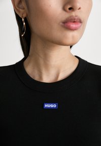 Crna rebrasta dukserica s plavim pravokutnim "HUGO" logom na prsima. Prikaz u krupnom planu s vidljivom kožom i zlatnom naušnicom.