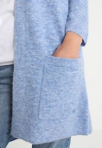 Osoba w jasnoniebieskim, puszystym kardiganie z ręką w przedniej kieszeni, w parze z niebieskimi dżinsami i białą koszulą.