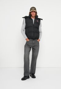 Melnā pūka veste ar augstu apkakli un rāvējslēdzēju, uzvilkta virs pelēka garām piedurknēm krekla. Modelis arī valkā pelēkus džinsus un melnas kurpes.
