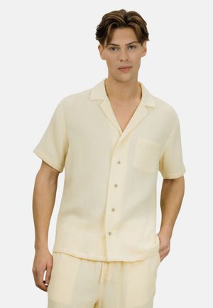 Ung mann med kort brunt hår iført en kremfarget kortermet skjorte med knapper og matchende løse bukser, stående med en hånd i lomma.