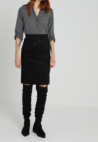 Svart mocka knälång kjol med dragsko, ihop med en knappad grå blus med prickar. Modellen bär slappa svarta knähöga stövlar.