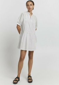 Eine Frau, die in einem weißen, kurzärmligen, oberschenkel-langen Kleid mit Spitzendetails steht und schwarze Plateausandalen trägt, vor einem schlichten weißen Hintergrund.