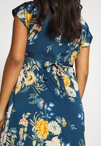 Nainen, jolla on päällään sininen mekko, jossa on oransseja, valkoisia ja vaaleanpunaisia kukkakuoseja. Mekko on sidottu takaa listalla, ja kuva näyttää hänen ylävartalonsa takaa.