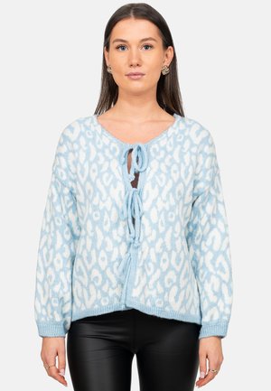 Woman wearing a light blue and white patterned knit cardigan with three front ties over black leather pants, standing against a plain background.
