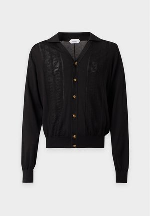 Jeune homme portant un cardigan noir boutonné par-dessus un t-shirt blanc, un pantalon large rayé et des baskets blanches et marron, debout les mains dans les poches.