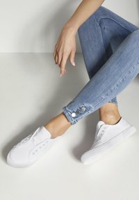 Weiße Leinenschuhe zum Hineinschlüpfen mit Gummisohlen, ausgestattet mit Metallösen. Kombiniert mit hellblauen Jeans aus Denim mit geknöpften Säumen.