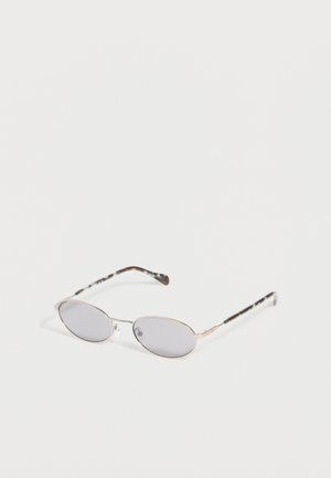 Occhiali da sole ovali con montatura argentata, lenti grigio chiaro e sottili aste decorate, posati su una superficie bianca.