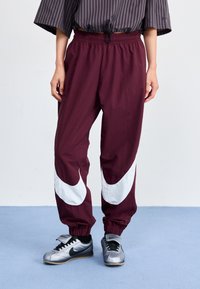 Bordeaux vindjakke bukser med hvite swoosh-design, elastisk midje og mansjetter. Kombinert med sølv Nike-sneakers.