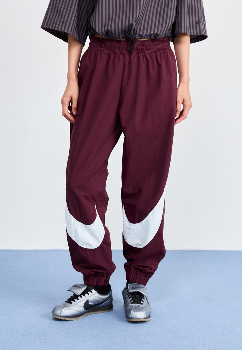 Bordeaux vindjakke bukser med hvite swoosh-design, elastisk midje og mansjetter. Kombinert med sølv Nike-sneakers.