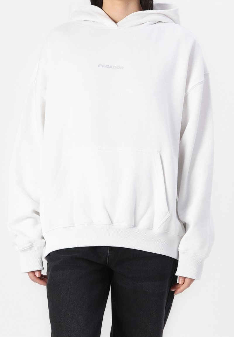 Sweat à capuche blanc surdimensionné avec une poche avant, des poignets et un ourlet côtelés, arborant un logo texte gris subtil "PEGADOR" sur la poitrine.