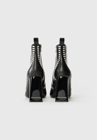 Bottines en cuir noir avec un talon bloc brillant, présentant des accents de couture blanche et une fermeture éclair à l'arrière.