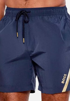 Homme portant un short de bain bleu marine avec un cordon noir et une bande diagonale blanche avec le logo BOSS sur la jambe gauche.
