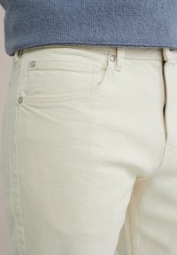 Pantalones de mezclilla beige claro con una superficie texturizada, que cuentan con un bolsillo frontal, botón de metal y cierre de cremallera. Combinados con una blusa de punto azul.