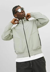 Light green zip-up jacket with high collar, two side pockets, and elastic hem. Worn over a white shirt and paired with oversized sunglasses.