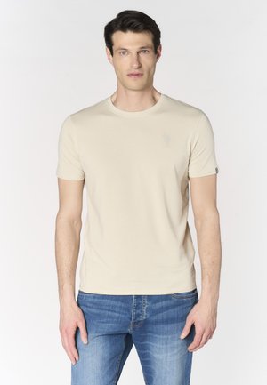 Uomo in piedi rivolto verso di noi che indossa una maglietta beige a maniche corte e jeans blu su uno sfondo bianco uniforme.