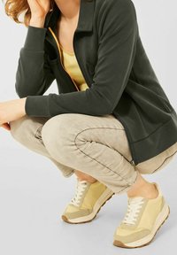 Giacca verde scuro con zip sopra una maglietta gialla, pantaloni beige chiaro e sneakers chiare con un mix di materiali testurizzati e accenti sottili.