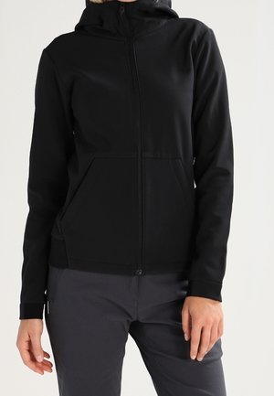 Persona vistiendo una sudadera negra con cremallera y bolsillos frontales, y pantalones gris oscuro, de pie contra un fondo claro y liso.