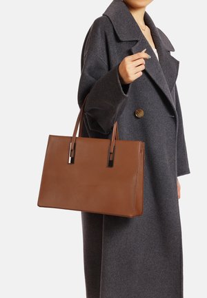 Borsa a tracolla in pelle marrone con un design strutturato, base piatta, due manici superiori e dettagli in metallo argentato. Indossata con un soprabito grigio.