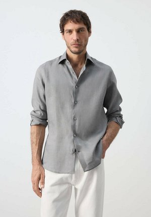 Mann mit dunklem Haar, der ein graues, geknöpftes Hemd mit hochgekrempelten Ärmeln und weiße Hosen vor einem schlichten hellen Hintergrund trägt.