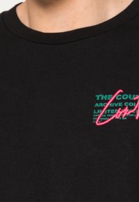 Camiseta de algodón negra con un gráfico de texto en verde y rosa en el pecho. Cuello redondo, mangas cortas y textura suave.