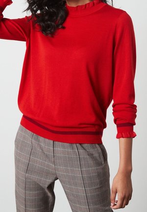 Femme portant un pull à manches longues rouge avec des poignets et un col volants, associé à un pantalon à carreaux gris sur un fond uni.