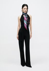 Black jumpsuit with a fitted silhouette, halter neckline, and a tie around the waist. Accented with a colorful patterned scarf.