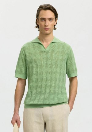 Bărbat tânăr purtând un polo tricotat cu mâneci scurte verde mentă cu modele de diamant și pantaloni bej deschis, ținând o curea de culoare deschisă.