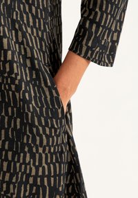 Robe noire avec un motif abstrait beige, des manches longues et une poche latérale. Le tissu a une apparence douce et texturée.