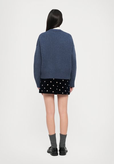 Masīva zila adīta džemperis ar apaļu kakla izgriezumu, ribotiem piedurkņu galiem un apakšmali; kombinēts ar melnu svārku ar sudraba rotājumiem.